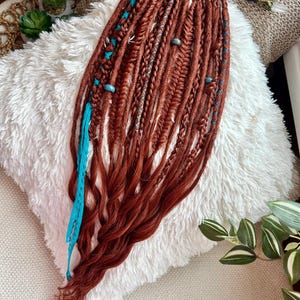 May include: Reddish-brown dreadlocks with turquoise accents and braids. The dreadlocks are arranged on a fluffy white surface. Some dreadlocks have turquoise beads and braids. A few strands of turquoise yarn are attached to the dreadlocks.
