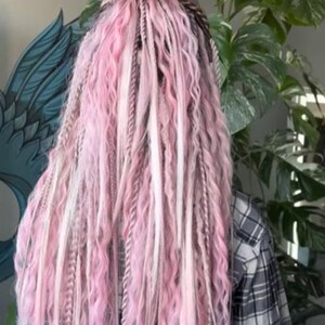 May include: A head of pink and white dreadlocks, styled in a bun. The dreadlocks are a mix of light pink, white, and gray, with a wavy texture. The person is wearing a plaid shirt.