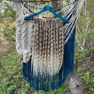 May include: A macrame wall hanging with a blue and cream color scheme, with a section of wavy, light brown and blonde hair extensions hanging from a teal hanger. The wall hanging is suspended from a wooden branch.