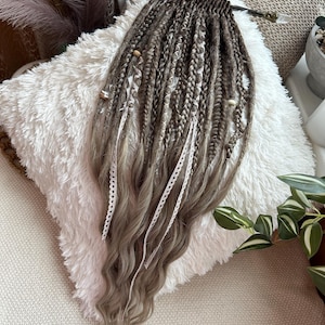 May include: A set of brown and gray dreadlocks with wavy ends, adorned with white lace and wooden beads. The dreadlocks are arranged on a fluffy white pillow, showcasing a natural hair extension style.