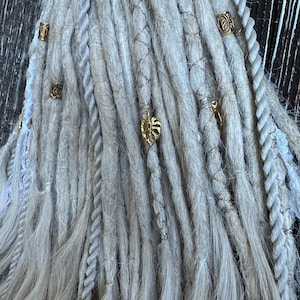 May include: Close-up of light gray dreadlocks with gold and silver accents. The dreadlocks are adorned with small gold beads and charms, including a leaf shape. Braided sections and silver thread add texture and detail to the hair.