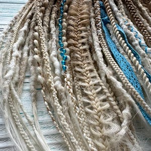 May include: A set of synthetic dreadlocks in various shades of white and brown, with some strands featuring blue beads and a blue braided cord. The dreadlocks are arranged in a fan shape on a white wooden surface.