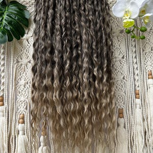 May include: A bundle of wavy, ombre hair extensions, transitioning from dark brown to a light blonde. The extensions are displayed against a macrame wall hanging with a wooden dowel and tassels. White orchids and green leaves are visible in the background.