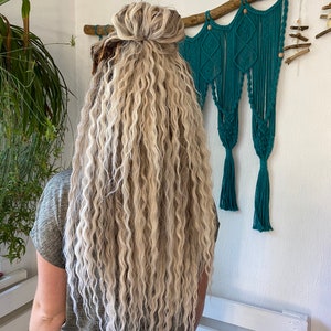 May include: A person with long, wavy, blonde and light brown hair styled in a bun. The hair cascades down the back. A teal macrame wall hanging and a white pallet are in the background.