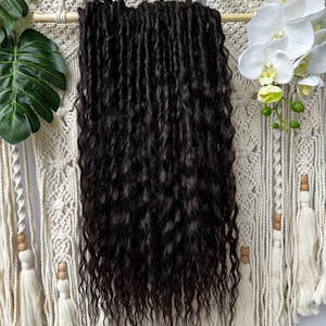 May include: Dark brown, wavy hair extensions suspended from a wooden rod. The extensions have a natural wave pattern. The background includes a macrame wall hanging with white tassels and decorative elements, plus green leaves and white orchids.