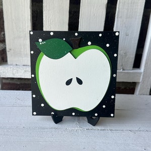 Puede incluir: Una manzana verde con un centro blanco y un tallo negro est&aacute; pintada sobre un fondo negro con lunares blancos. La manzana est&aacute; sobre un peque&ntilde;o caballete de madera.