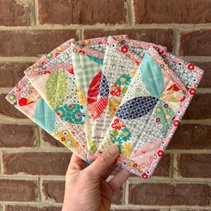 Puede incluir: Un abanico de posavasos de tela acolchada y colorida. Cada posavasos presenta un diseño de patchwork único con patrones florales y geométricos en tonos rosa, azul, verde y rojo. Los bordes están acabados con un ribete rosa.