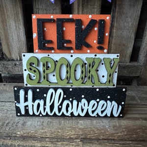 May include: Three wooden blocks stacked on top of each other. The top block is orange with white polka dots and says "EEK!" in black letters. The middle block is white with black polka dots and says "SPOOKY" in green letters. The bottom block is black with white polka dots and says "Halloween" in white letters.