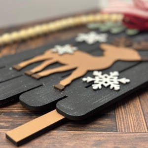 Puede incluir: Un letrero de Navidad de madera con fondo negro y una silueta de reno marr&oacute;n. El letrero est&aacute; decorado con copos de nieve blancos y tiene un palo de madera marr&oacute;n unido a la parte inferior.