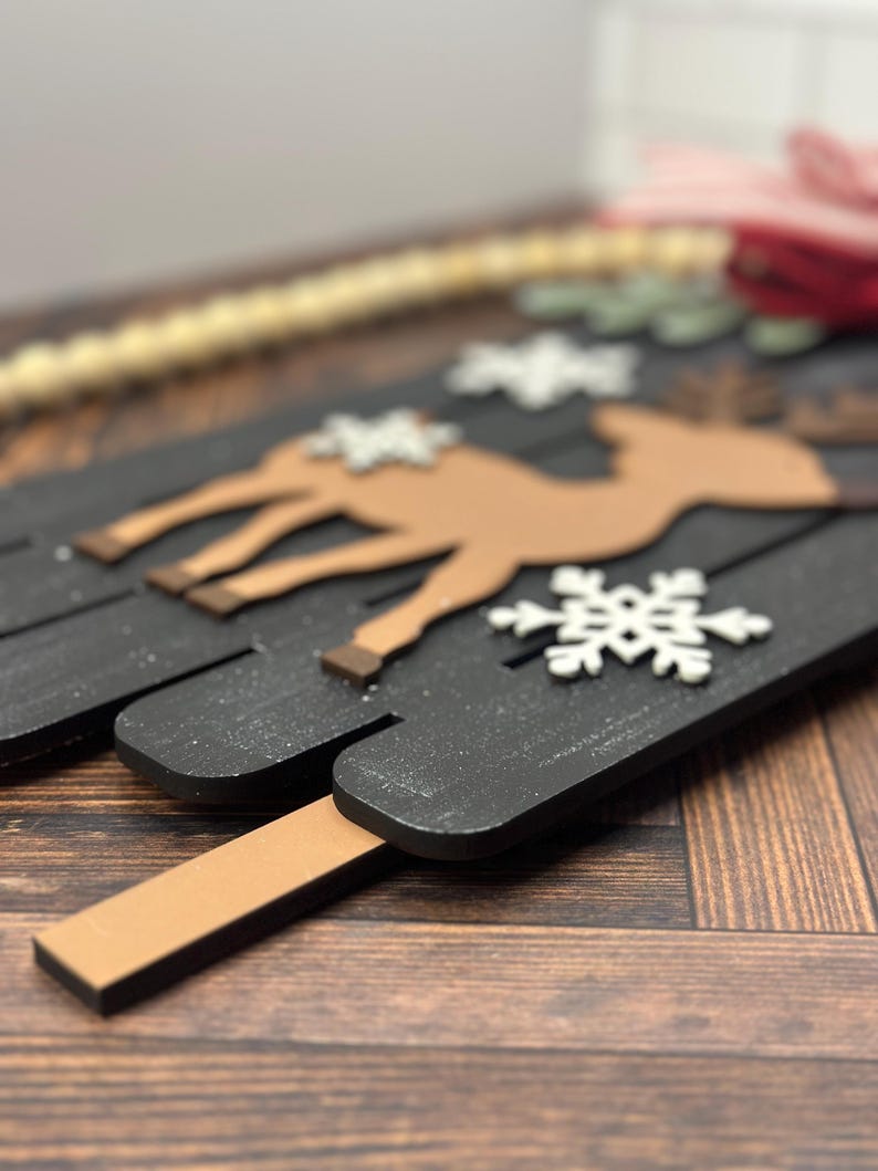 Puede incluir: Un letrero de Navidad de madera con fondo negro y una silueta de reno marr&oacute;n. El letrero est&aacute; decorado con copos de nieve blancos y tiene un palo de madera marr&oacute;n unido a la parte inferior.