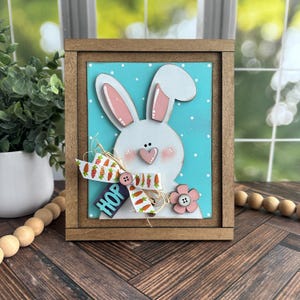 May include: A wooden framed sign with a white bunny face on a turquoise background with white polka dots. The bunny has pink ears and a pink heart nose. The sign says "HOP" in blue letters and has a carrot and flower embellishment.