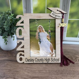 Puede incluir: Un marco de fotos de graduación de madera con el año "2026" recortado en el lado izquierdo. El marco contiene una foto de una persona en un columpio. Una gorra de graduación y una borla están adjuntas, con el nombre "Alyssa" y el año "2026" en la gorra.