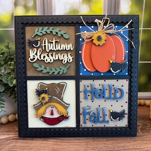 May include: A square, black-framed wooden sign with four decorative panels. The panels feature autumn-themed designs, including a pumpkin, scarecrow, and the phrases "Autumn Blessings" and "Hello Fall." The sign is adorned with small black birds and floral accents.