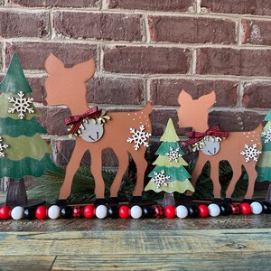 May include: A set of three wooden Christmas decorations featuring a deer, a tree, and a bell. The deer are brown with red and white bows. The trees are green with white snowflakes. The decorations are arranged in a row on a wooden surface.