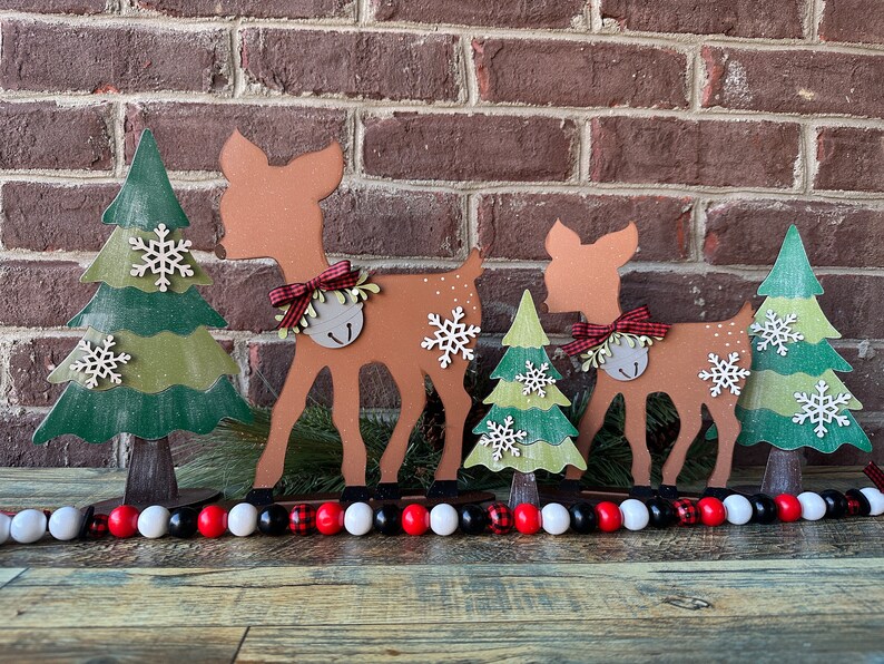 May include: A set of three wooden Christmas decorations featuring a deer, a tree, and a bell. The deer are brown with red and white bows. The trees are green with white snowflakes. The decorations are arranged in a row on a wooden surface.