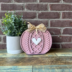 May include: A pink wooden pumpkin with a white heart cutout and a burlap bow. The pumpkin has a white polka dot pattern and a lattice design.