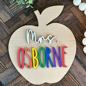 May include: A wooden apple-shaped sign with the text "Mrs. OSBORNE" in rainbow letters. The letters are attached to the sign with a white background.