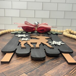 Puede incluir: Una decoraci&oacute;n de trineo de Navidad de madera con un reno marr&oacute;n, copos de nieve blancos y un lazo rojo y blanco a rayas con una campana.