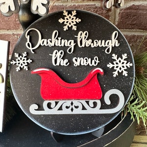 May include: Black and white round wooden sign with the text "Dashing through the snow" and a red sleigh with white trim. The sign has a glittery finish.