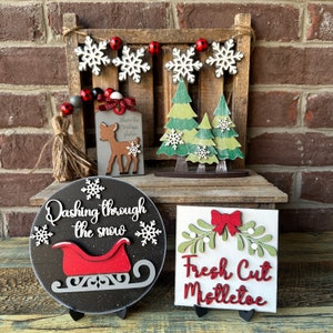 May include: A set of four wooden Christmas decorations. The first decoration is a round sign with the text "Dashing through the snow" and a red sleigh. The second decoration is a square sign with the text "Fresh Cut Mistletoe" and a green sprig of mistletoe with a red bow. The third decoration is a rectangular sign with the text "Where the treetops glisten" and a brown deer. The fourth decoration is a set of two green Christmas trees with white snowflakes.