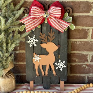 Puede incluir: Una decoraci&oacute;n de trineo de Navidad de madera con un lazo rojo y blanco a rayas, una campana y un recorte de reno marr&oacute;n. El trineo est&aacute; pintado de negro y tiene un acabado desgastado.