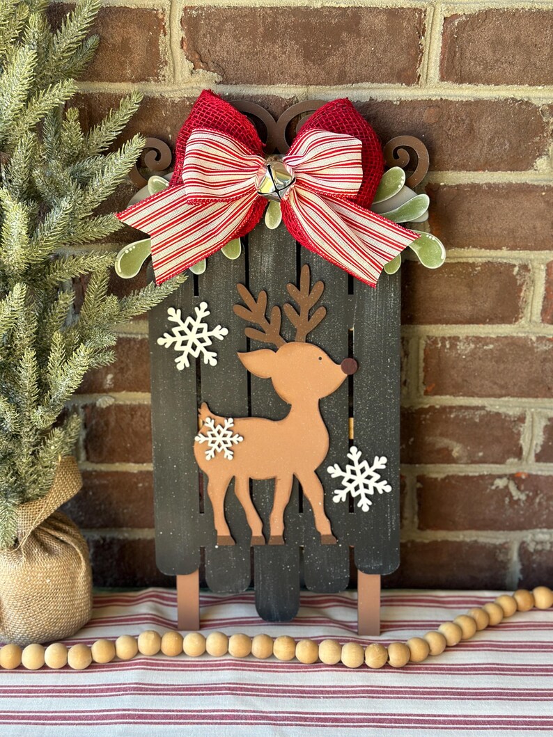 Puede incluir: Una decoraci&oacute;n de trineo de Navidad de madera con un lazo rojo y blanco a rayas, una campana y un recorte de reno marr&oacute;n. El trineo est&aacute; pintado de negro y tiene un acabado desgastado.