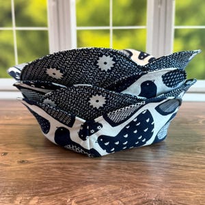 May include: A stack of three fabric bowl cozies. The cozies feature a navy blue and white bird and floral pattern. The interior of the cozies has a navy blue and white geometric pattern. The cozies are sitting on a wooden surface.
