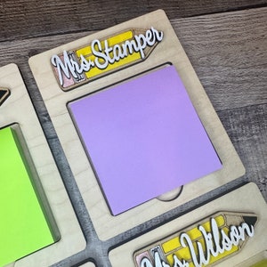 May include: A wooden desk organizer with two slots for sticky notes. The top slot has a purple sticky note and a yellow pencil with the text "Mrs. Stamper". The bottom slot has a yellow pencil with the text "Mrs. Wilson".