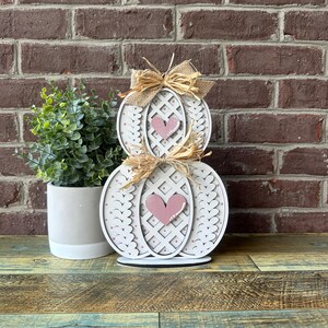 May include: A white wooden pumpkin with a lattice design and pink hearts. The pumpkin is decorated with a burlap bow and is stacked on top of another white pumpkin with a similar design.