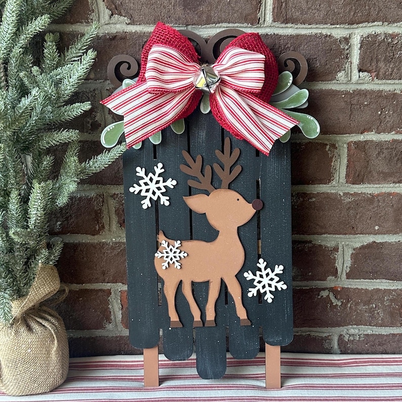 Puede incluir: Una decoraci&oacute;n navide&ntilde;a de madera en forma de trineo de reno con un lazo rojo y blanco a rayas. El trineo est&aacute; pintado de negro y tiene un reno marr&oacute;n con copos de nieve blancos.