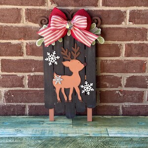 Puede incluir: Un trineo de Navidad de madera con un lazo rojo y blanco a rayas, una campana y un recorte de reno marr&oacute;n. El trineo est&aacute; decorado con copos de nieve blancos.