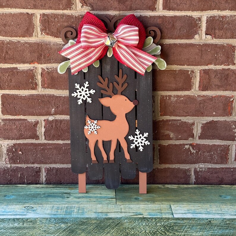 Puede incluir: Un trineo de Navidad de madera con un lazo rojo y blanco a rayas, una campana y un recorte de reno marr&oacute;n. El trineo est&aacute; decorado con copos de nieve blancos.