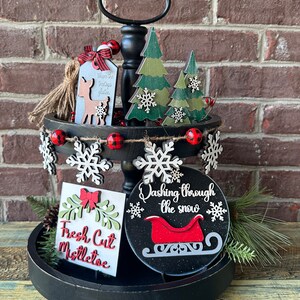 May include: A two-tier black tiered tray decorated for Christmas. The top tier features a wooden deer with a tag that says "Where the trees whisper" and two green wooden trees with snowflakes. The bottom tier features a wooden sign that says "Fresh Cut Mistletoe", a round wooden sign that says "Dashing through the snow" with a red sleigh, and a small pine sprig. The tray is decorated with red and black buffalo check ribbon, white snowflakes, and red and white beads.