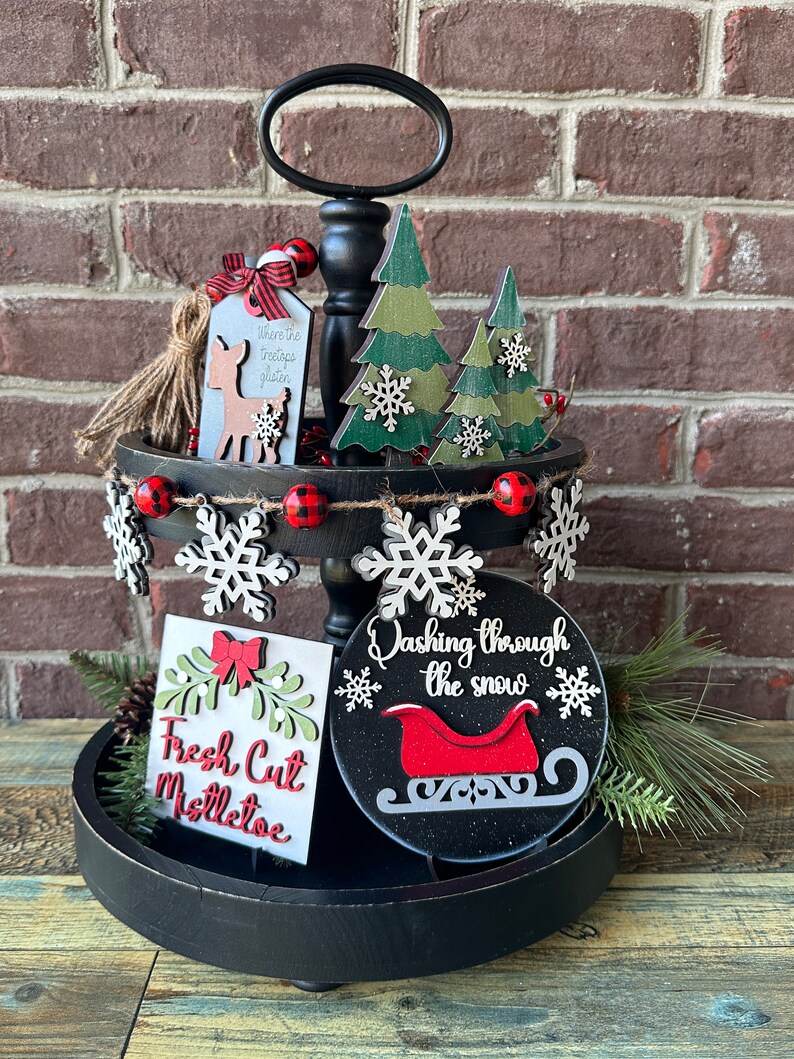 May include: A two-tier black tiered tray decorated for Christmas. The top tier features a wooden deer with a tag that says "Where the trees whisper" and two green wooden trees with snowflakes. The bottom tier features a wooden sign that says "Fresh Cut Mistletoe", a round wooden sign that says "Dashing through the snow" with a red sleigh, and a small pine sprig. The tray is decorated with red and black buffalo check ribbon, white snowflakes, and red and white beads.