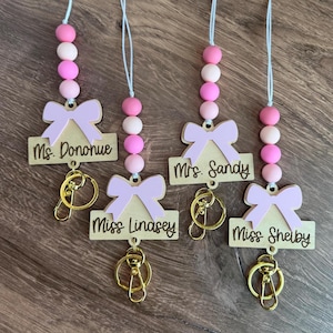 May include: Four teacher lanyards with pink and white beads. Each lanyard has a wooden tag with a light pink bow and a name, plus a gold-coloured keychain. The names on the tags are Ms. Donohue, Mrs. Sandy, Miss Linasey, and Miss Shelby.
