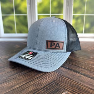 May include: Gray trucker hat with a black mesh back and a curved brim. The hat features a brown leather patch with the letters "PA" in black. The Richardson 112 adjustable hat has a structured front and a snapback closure.