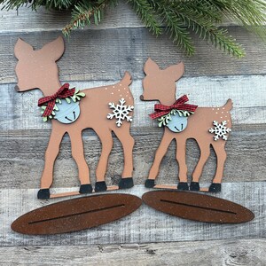 May include: Two wooden deer ornaments painted brown, with red and white checked bows and silver bells. The deer are standing on brown wooden bases.