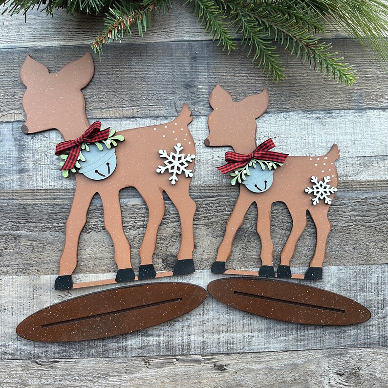 May include: Two wooden deer ornaments painted brown, with red and white checked bows and silver bells. The deer are standing on brown wooden bases.
