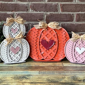 May include: Three wooden pumpkins decorated with hearts and a woven pattern. The pumpkins are painted in shades of white, pink, and red.