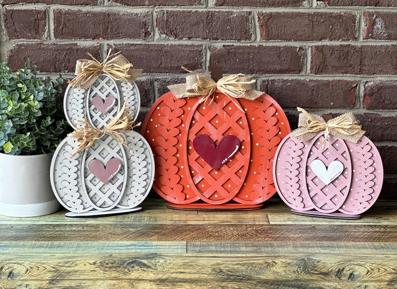 May include: Three wooden pumpkins decorated with hearts and a woven pattern. The pumpkins are painted in shades of white, pink, and red.