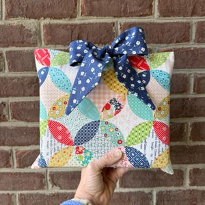 May include: A colorful, handmade pillow with a patchwork design featuring red, blue, green, and yellow fabrics. The pillow is adorned with a navy blue bow with a white floral pattern. The pillow is held in a person's hand.