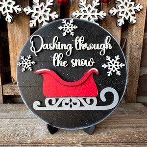 May include: A black wooden round sign with white snowflakes and the text "Dashing through the snow" in white script. A red sleigh with a white trim is attached to the sign.