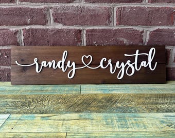 May include: A wooden sign with the names "Randy" and "Crystal" written in white cursive lettering with a heart symbol between the names.