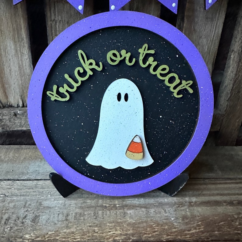 May include: Purple and black round sign with the words "trick or treat" in gold letters. A white ghost with a candy corn is in the center of the sign.