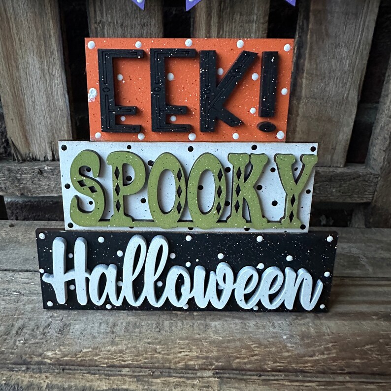 May include: Three wooden blocks stacked on top of each other. The top block is orange with white polka dots and says "EEK!" in black letters. The middle block is white with black polka dots and says "SPOOKY" in green letters. The bottom block is black with white polka dots and says "Halloween" in white letters.