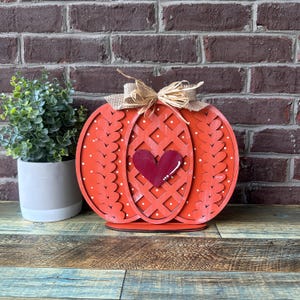 May include: A red wooden pumpkin with white polka dots and a woven heart design. The pumpkin has a burlap bow and a small red heart in the centre.