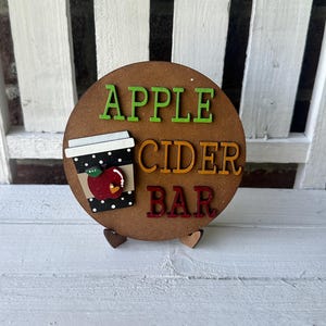 Puede incluir: Un letrero redondo de madera con las palabras "Apple Cider Bar" en letras de colores. Una taza negra y blanca con una manzana roja y lunares blancos est&aacute; en el letrero.