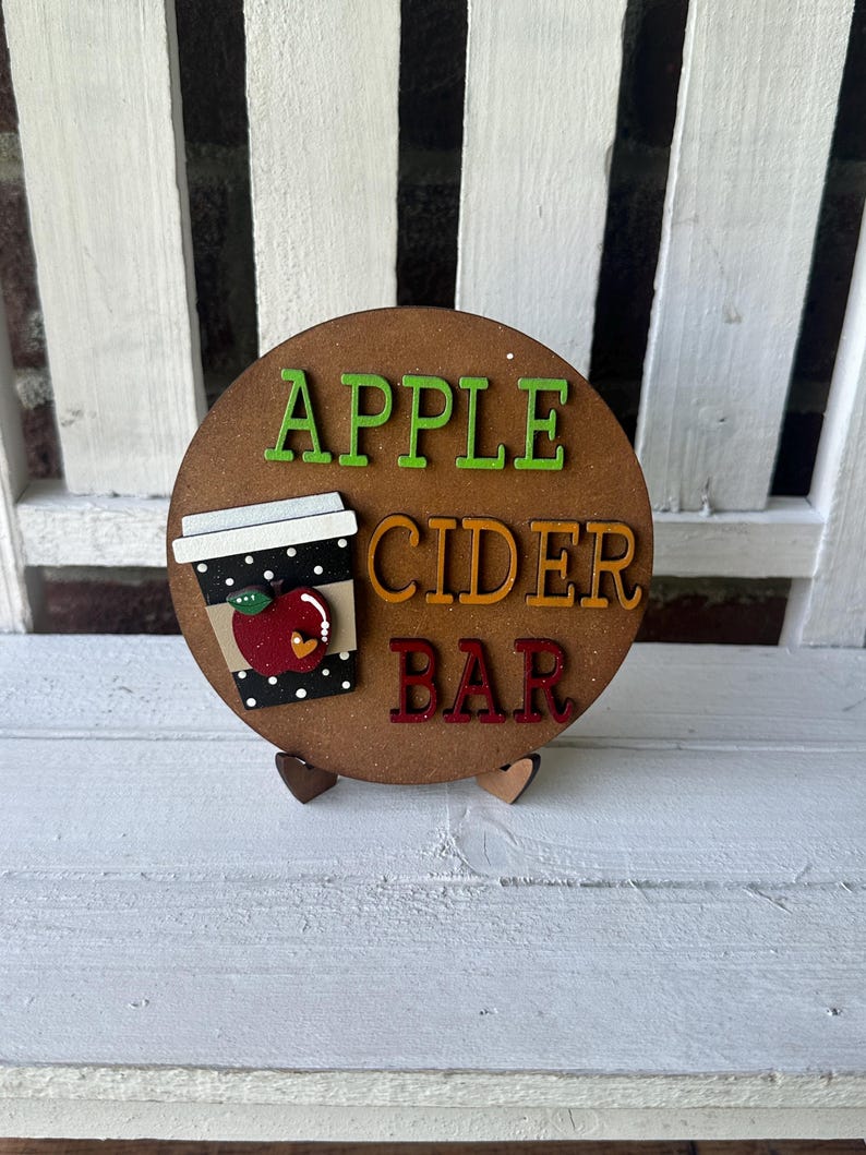 Puede incluir: Un letrero redondo de madera con las palabras "Apple Cider Bar" en letras de colores. Una taza negra y blanca con una manzana roja y lunares blancos est&aacute; en el letrero.