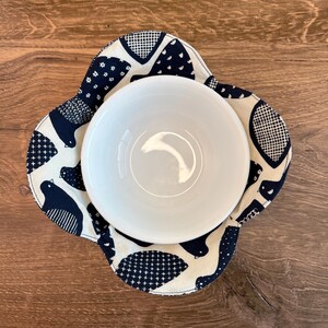 May include: A white ceramic bowl sits inside a navy blue and cream-colored bowl cozy. The cozy features a repeating pattern of birds and geometric shapes. The bowl and cozy are on a wooden surface.