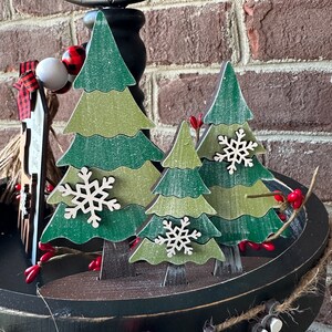 May include: Three wooden Christmas trees with a distressed white paint finish. The trees are decorated with white snowflakes and are arranged on a black tiered tray.